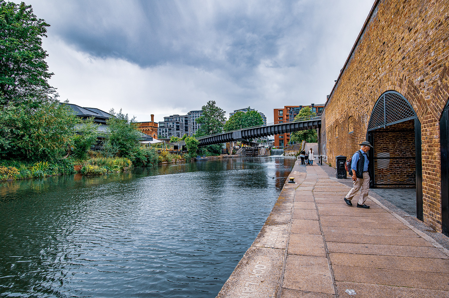 The image depicts a serene canal scene with a person walking along a brick-paved path beside the water. The area is surrounded by lush greenery and modern buildings, with a pedestrian bridge in the background. The sky is overcast, suggesting a cloudy day. The scene captures a blend of natural and urban elements, creating a peaceful and picturesque environment.