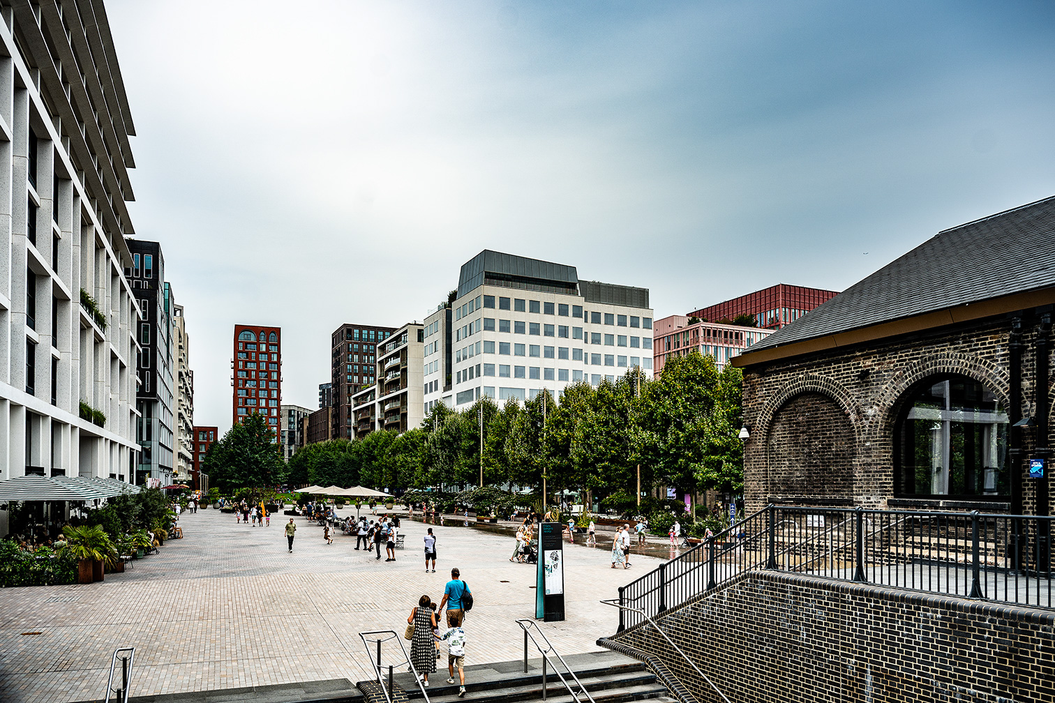 The image depicts a bustling urban plaza with modern buildings, a mix of architectural styles, and people walking and relaxing in the open space. The area is lined with trees and outdoor seating, creating a lively and inviting atmosphere.