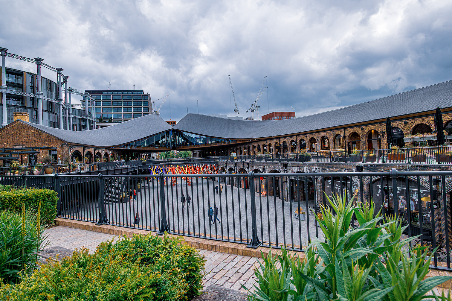 The image depicts a bustling market area with a mix of modern and historic architecture. The central building features arched windows and a grey roof, while modern buildings and cranes are visible in the background. The area is enclosed by a black fence, with greenery and plants in the foreground. People are walking around, indicating an active and lively environment.