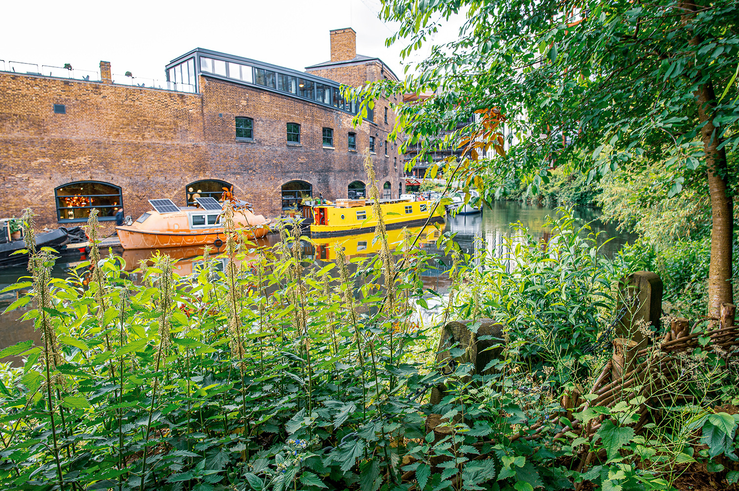 The image depicts a serene canal scene with brick buildings on one side and lush greenery on the other. Two boats are moored along the canal, with one boat being orange and the other yellow. The buildings have a rustic, industrial look with large windows and a brick facade. The greenery is dense with various plants and trees, adding a natural touch to the urban setting