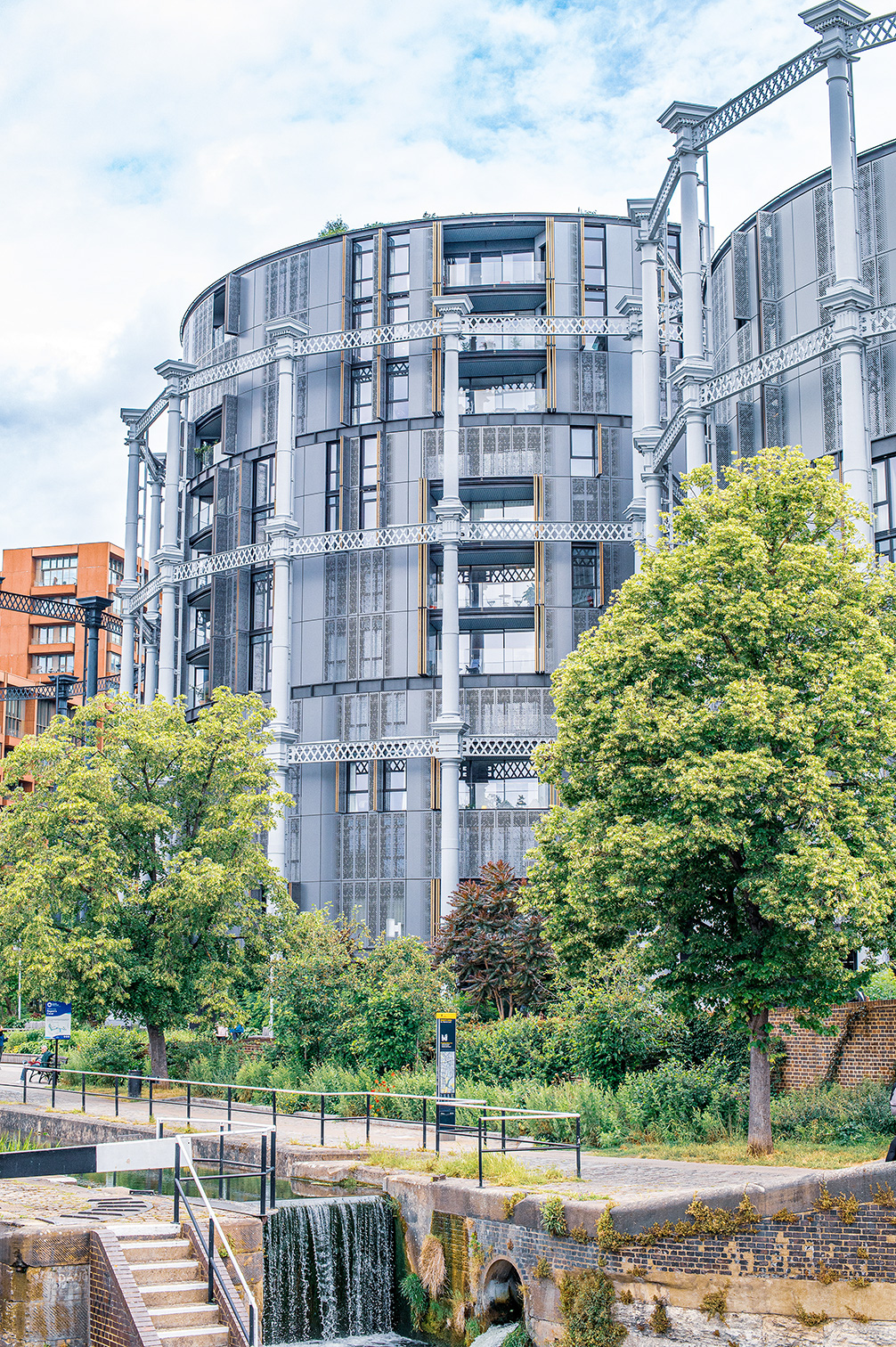 The image depicts a modern, multi-story building with a distinctive architectural design, featuring large glass windows and intricate metal railings. The building is surrounded by lush greenery and trees, with a small waterfall and a pathway leading up to it. The sky is partly cloudy, and the overall scene suggests a blend of urban and natural elements.