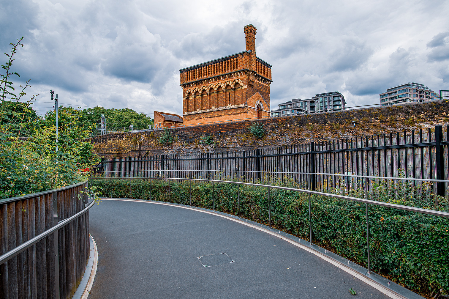 The image depicts an old brick building with a distinctive architectural style, featuring arched windows and a prominent chimney. The building is situated on elevated ground, with a tall brick wall covered in ivy and other vegetation. In the foreground, there is a curved pathway with metal railings, leading towards the building. Modern residential buildings are visible in the background, contrasting with the historical structure. The sky is overcast, adding a dramatic effect to the scene.