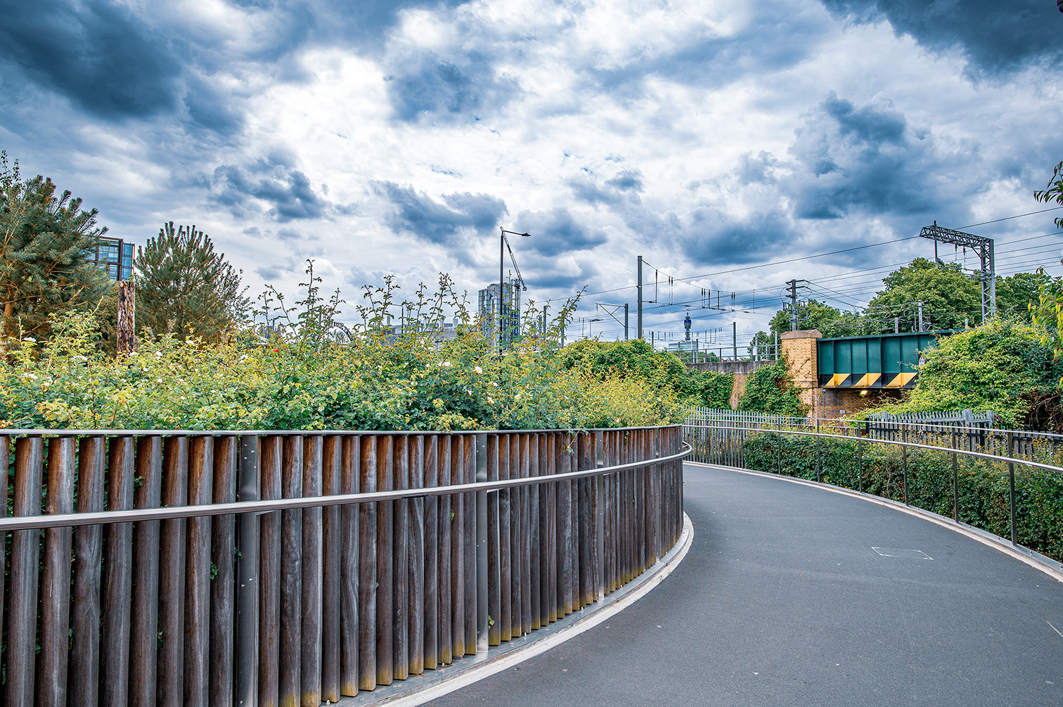 The image depicts a curved pathway with a metal railing on the left side, leading towards a bridge. The path is surrounded by lush greenery and vegetation, with tall plants and trees on both sides. In the background, there are buildings, power lines, and a cloudy sky, suggesting an urban setting with natural elements.