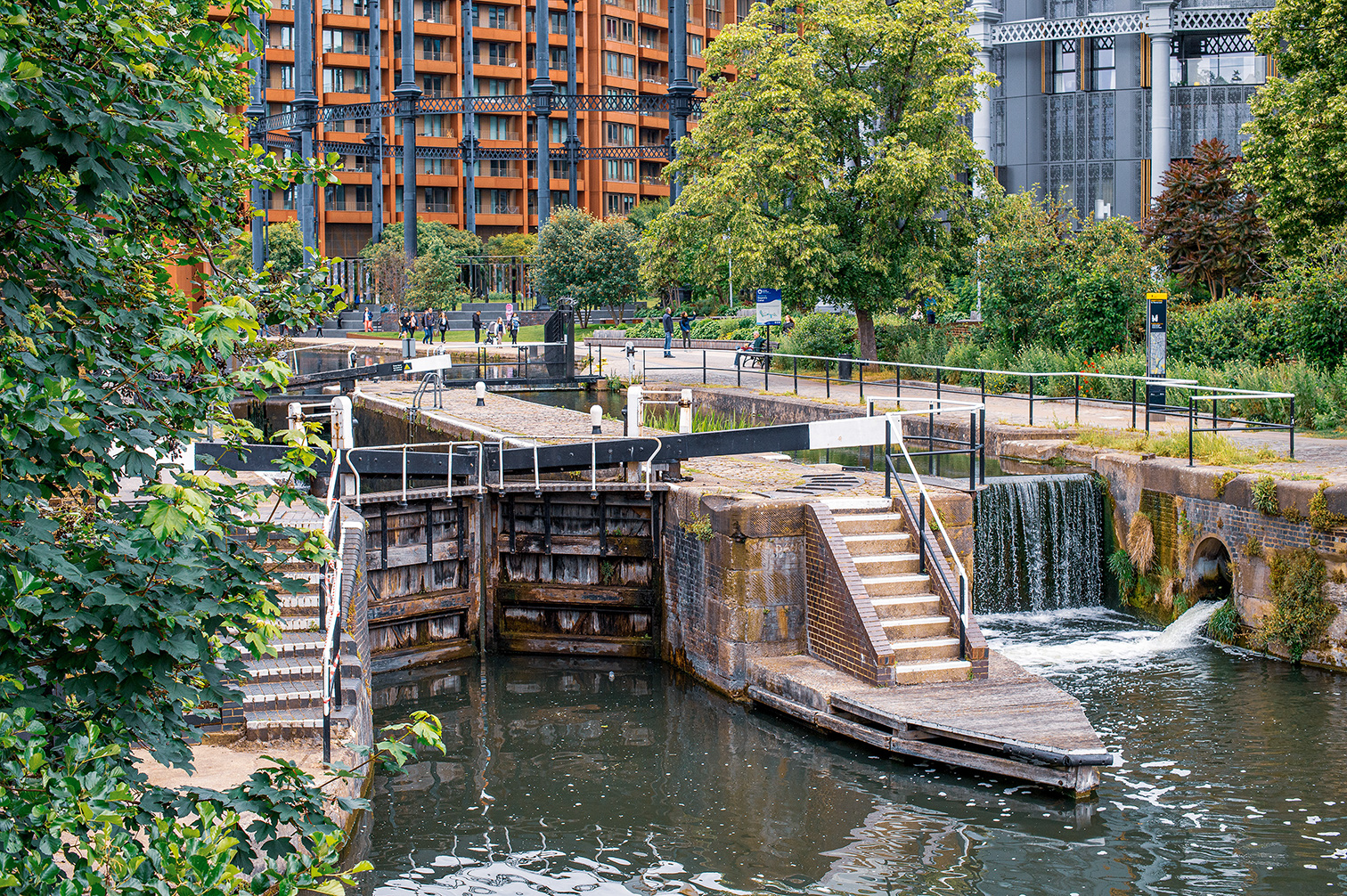 The image depicts a serene urban scene featuring a canal with a lock system, surrounded by lush greenery and modern apartment buildings in the background. People are seen walking along a path adjacent to the canal, enjoying the peaceful environment. The lock gates are open, allowing water to flow through, and there are steps leading up to the path from the lock structure. The overall atmosphere is calm and inviting, blending natural elements with urban living.