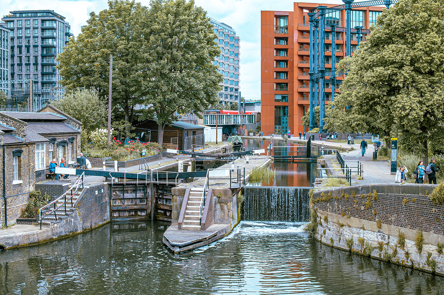 The image depicts a canal lock system in an urban environment. The lock is open, allowing water to flow through, and is surrounded by brick structures and greenery. Modern high-rise buildings are visible in the background, while people are seen walking and relaxing along the canal paths. The scene suggests a blend of natural and urban elements, with the canal serving as a recreational area for residents.