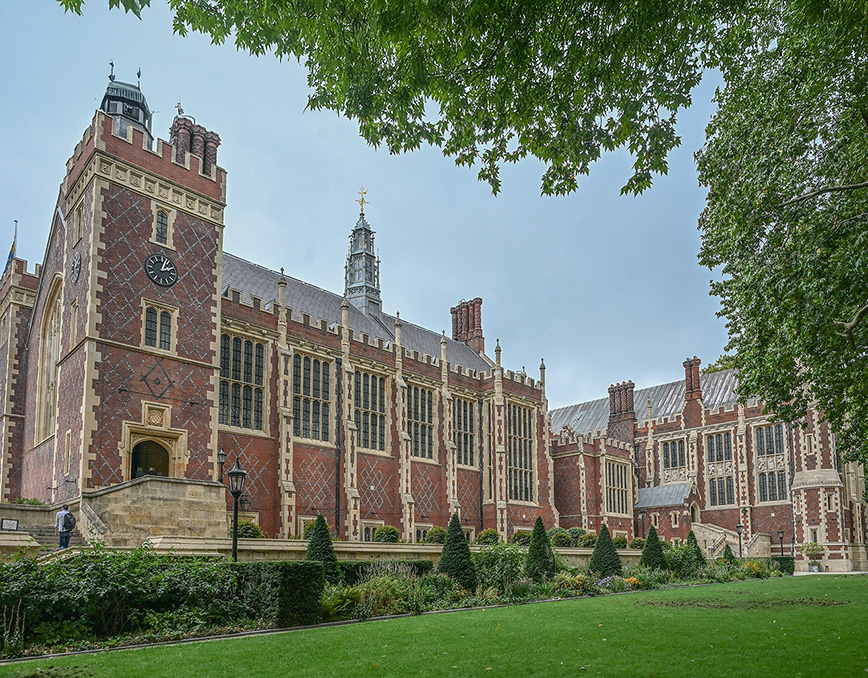 The image depicts a large, historic building with intricate architectural details, featuring tall towers, a clock, and numerous large windows. The structure is made of red and brown bricks with decorative stone accents. The building is surrounded by well-maintained gardens with green lawns, shrubs, and trees, suggesting it could be part of a university or a significant public institution.