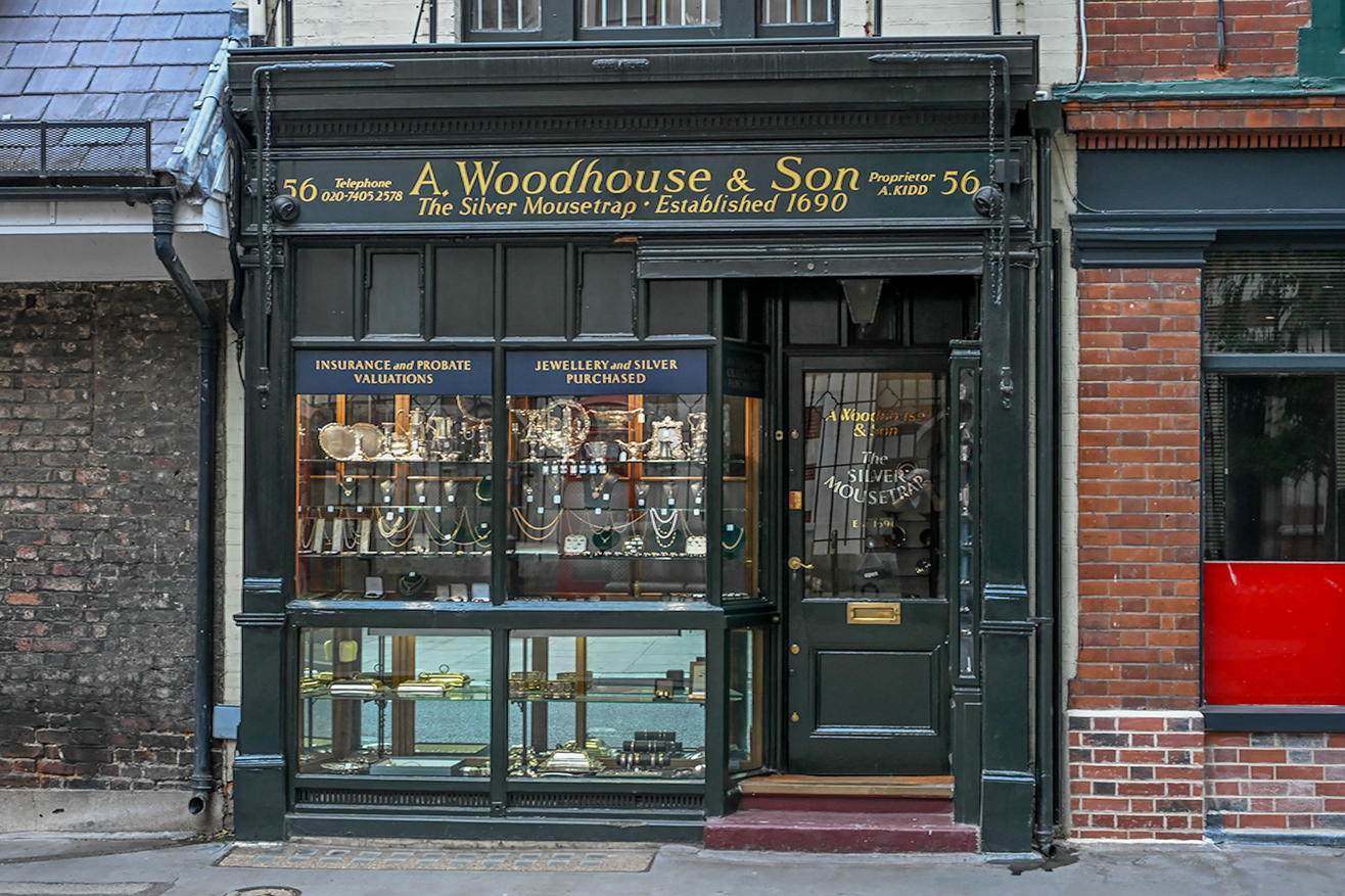 The image depicts the storefront of 'A. Woodhouse & Son,' a shop specializing in jewelry and silver, established in 1690. The shop offers services such as insurance and probate valuations. The facade features a traditional design with a glass display showcasing various silver items and jewelry. The address is 56, and the telephone number provided is 020-74052578. The proprietor is A. Kidd.