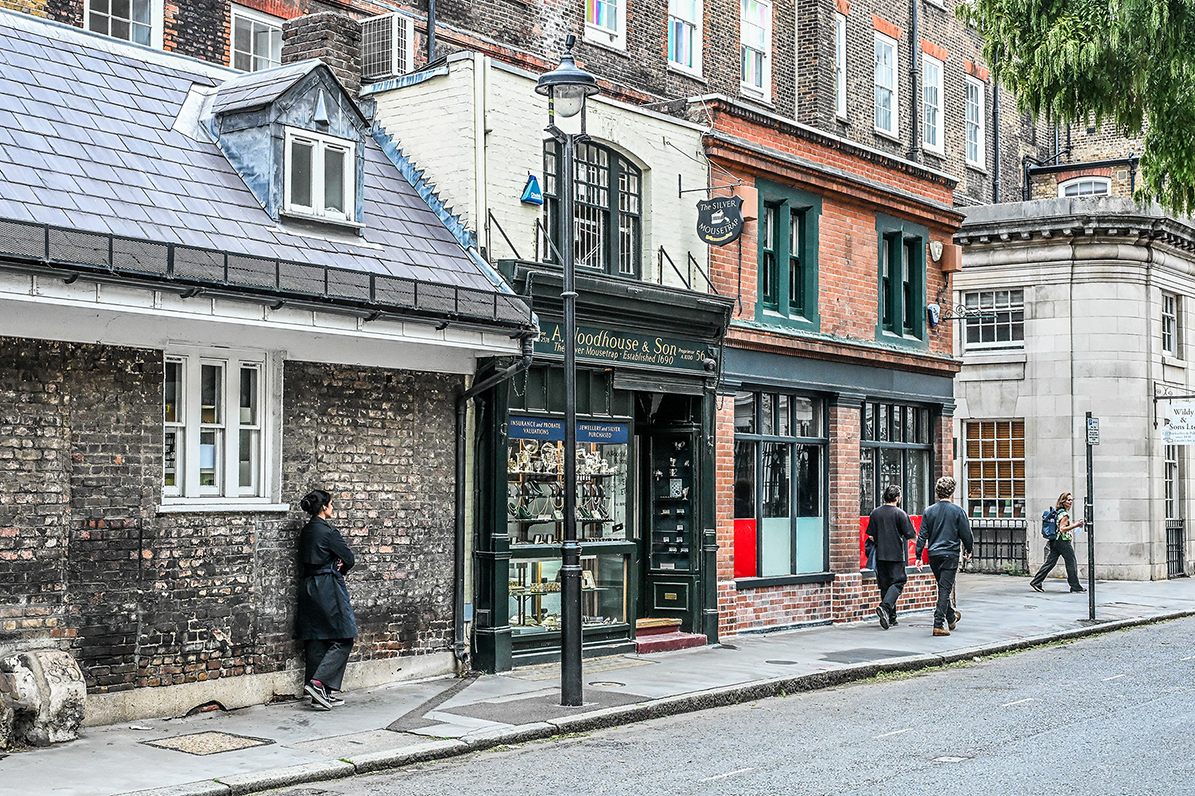 The image depicts a street scene in what appears to be a historic or older part of a city. There are several people walking along the sidewalk, and a woman is standing near a building. The buildings have a mix of brick and stone facades, with some featuring large windows and others smaller, more traditional windows. One of the buildings houses a shop named 'A. Goodhouse & Son,' which seems to sell jewelry and antiques. The street is relatively narrow and lined with traditional street lamps.