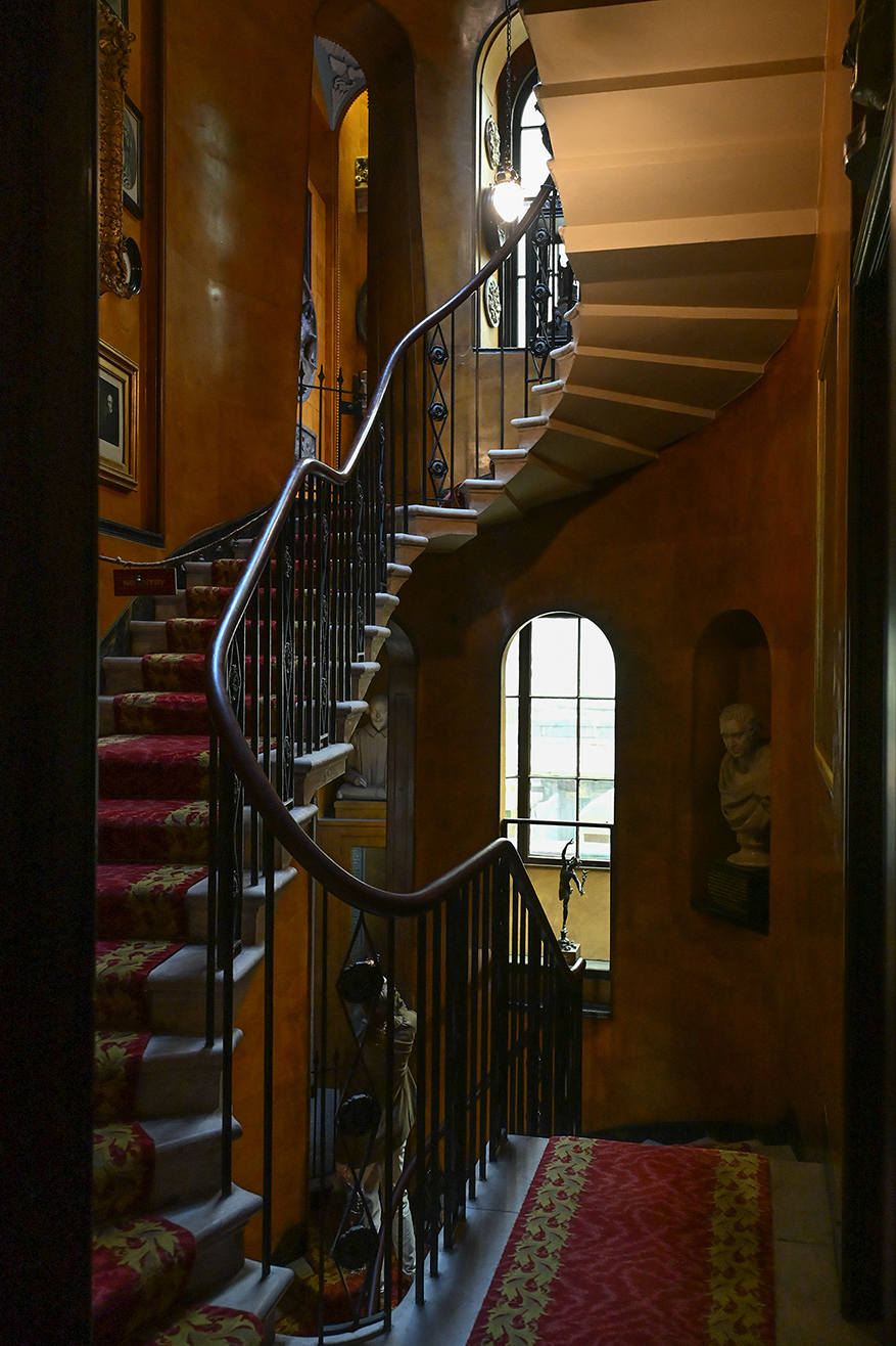 The image depicts an elegant, spiral staircase in a well-lit, ornately decorated interior. The staircase features a wrought iron railing and is adorned with a red and gold patterned carpet. The walls are decorated with framed artworks and a bust statue is placed in a niche by a window, allowing natural light to illuminate the space