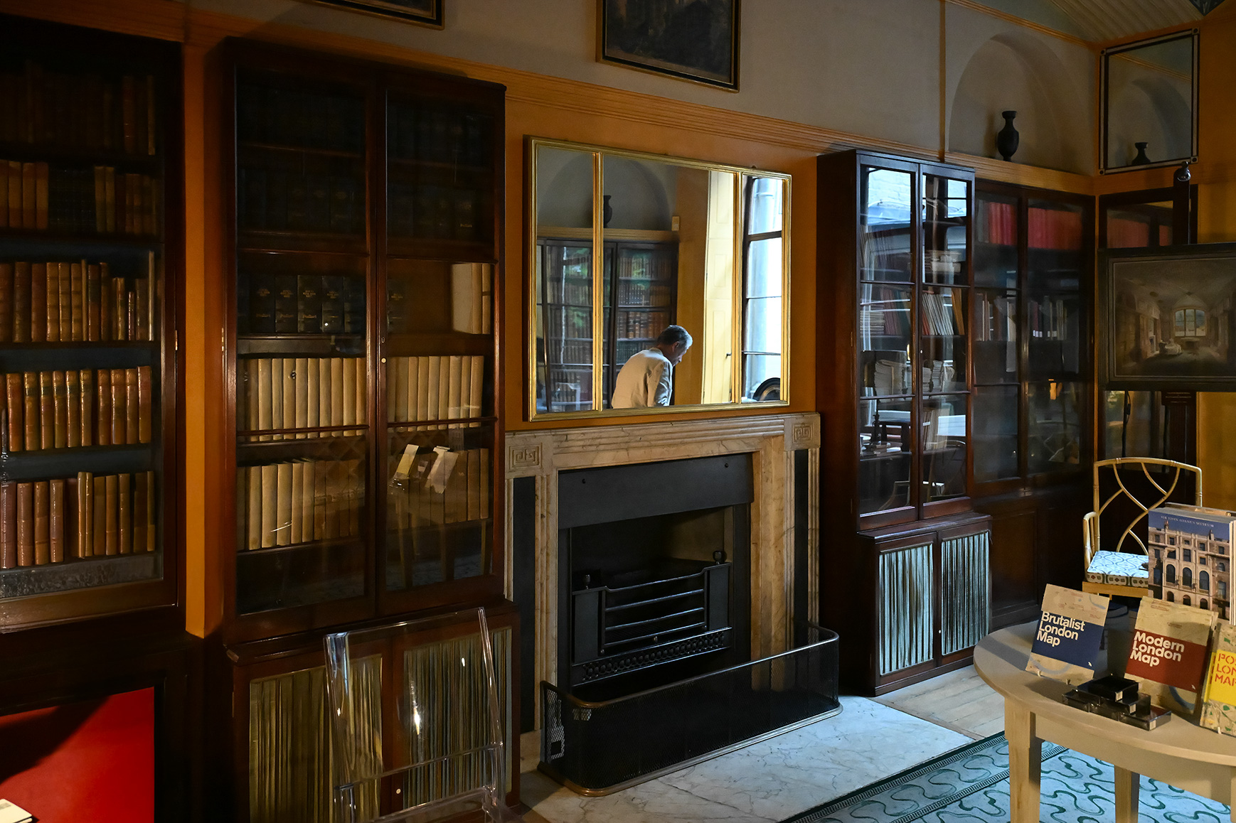 The image depicts a cozy, well-lit room with wooden bookshelves filled with books and glass-fronted cabinets. The room features a fireplace with a wooden mantel, and a person is seen through a window, leaning in to look at something. There are various items on display, including maps and other reading materials on a small table in the foreground. The overall ambiance suggests a quiet, scholarly environment, possibly a library or study.