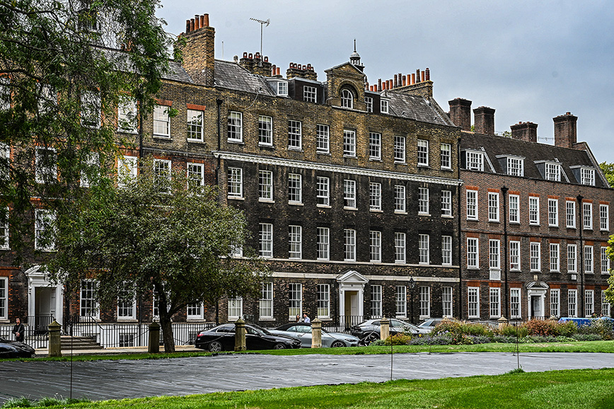 The image depicts a row of traditional, multi-story buildings with a classic architectural style, featuring brick facades and numerous windows. The buildings are set back from the street, with a small parking area and a grassy area in front. The scene is framed by trees on the left side, and the overall atmosphere is calm and orderly.