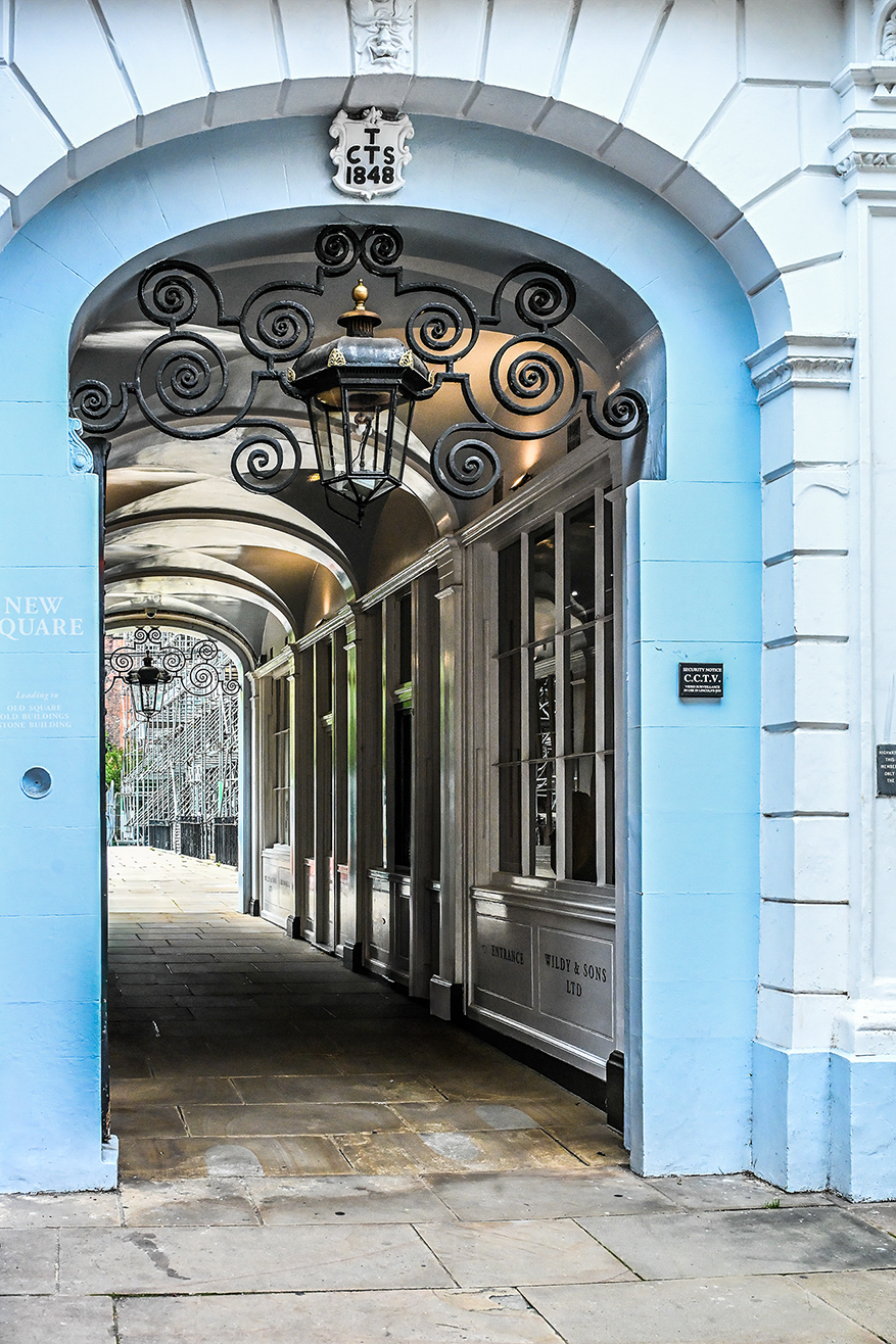 The image depicts an entrance to a building with the name 'Wild & Sons Ltd.' The entrance is adorned with ornate, vintage-style ironwork and a lantern. The archway above the entrance bears the inscription 'T CTS 1848' and a sign indicating CCTV surveillance. The building is part of 'New Square' and the entrance is flanked by light blue walls with white accents. The pathway leading into the entrance is paved with ston