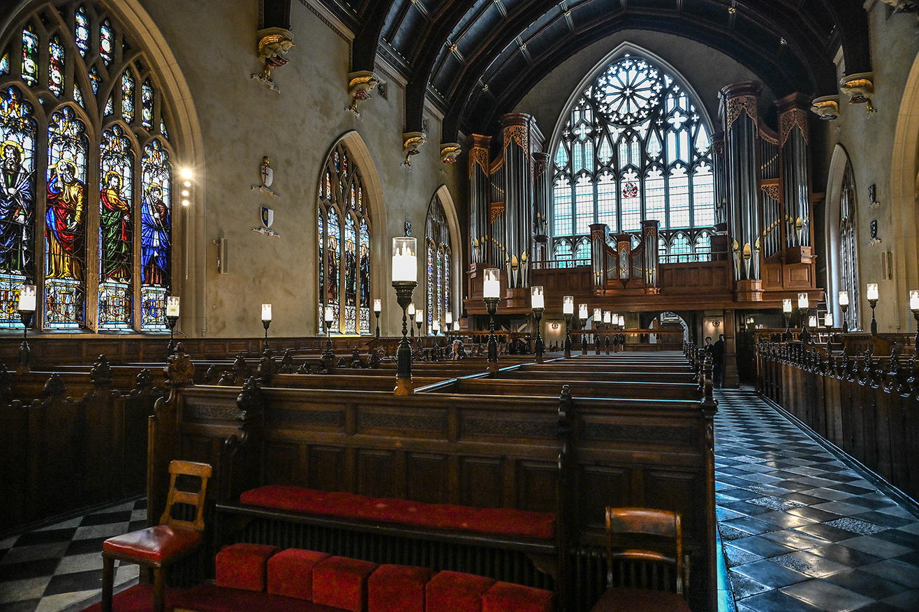 The image depicts the interior of a grand, Gothic-style church. The scene features intricate stained glass windows, wooden pews with red cushions, and a large pipe organ. The architecture includes pointed arches, ribbed vaults, and detailed stone carvings. The lighting is dim, with natural light streaming through the stained glass, creating a serene and reverent atmosphere. The overall setting is one of historical and architectural significance, likely used for worship and reflection.