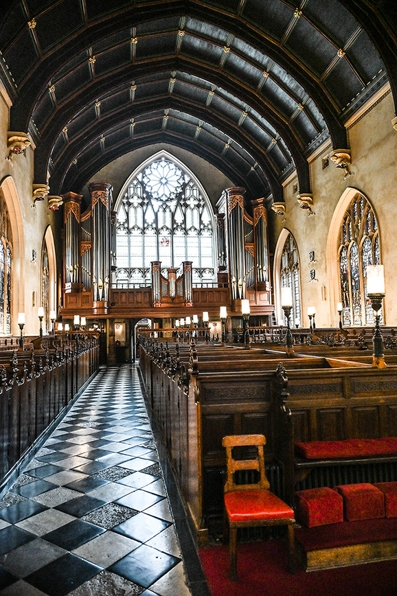 The image depicts the interior of a grand, Gothic-style church. The viewer's perspective is from the center aisle, looking towards the altar. The ceiling features intricate, ribbed vaulting, and the walls are adorned with tall, arched windows filled with stained glass. Wooden pews line both sides of the aisle, and a large, ornate pipe organ is visible at the front of the church. The overall atmosphere is one of solemnity and reverence.