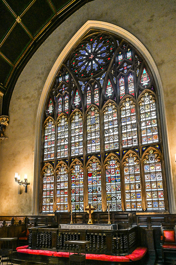 The image depicts an interior view of a church, showcasing a large, ornate stained-glass window behind the altar. The window features intricate designs and vibrant colors, with various religious symbols and figures. The altar area is adorned with wooden furniture, including pews with red cushions and a central altar table with a cross. The overall atmosphere is serene and reverent, typical of a place of worship.