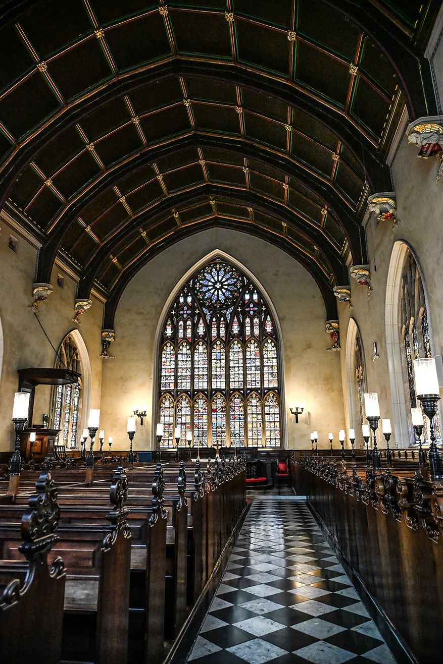 The image depicts the interior of a grand, traditional church. The view is down the central aisle, flanked by wooden pews with ornate ends. The ceiling features a ribbed vault with gold detailing, and the walls are adorned with large, colorful stained-glass windows that allow natural light to filter into the space. The floor is tiled in a black and white checkerboard pattern, and the overall atmosphere is one of solemnity and reverence.