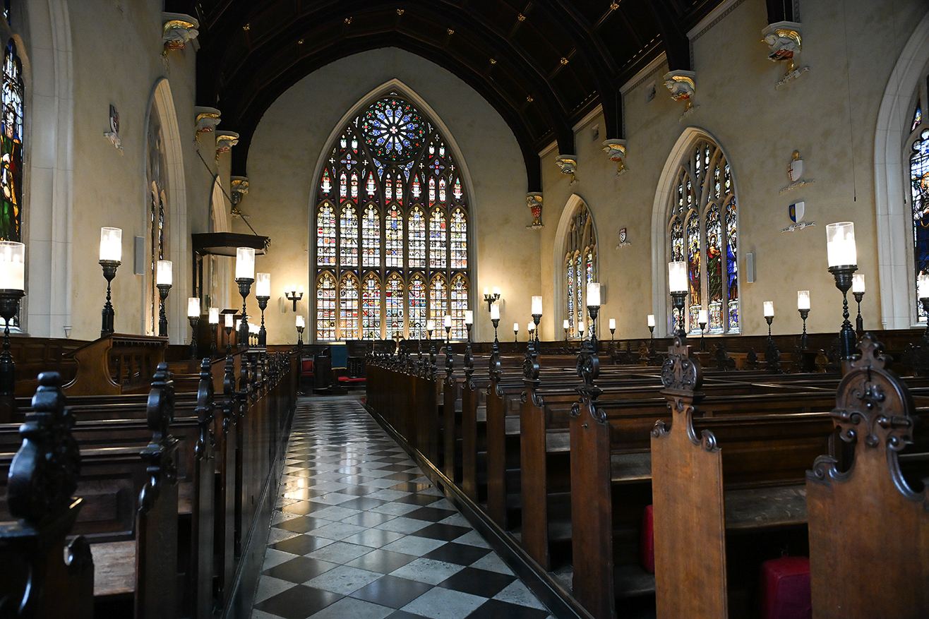 The image depicts the interior of a church, showcasing rows of wooden pews with black metal candleholders, leading up to an altar. The space is illuminated by wall-mounted lights and natural light streaming through large, intricate stained-glass windows. The architecture features high, arched ceilings and detailed decorations, creating a serene and reverent atmosphere.