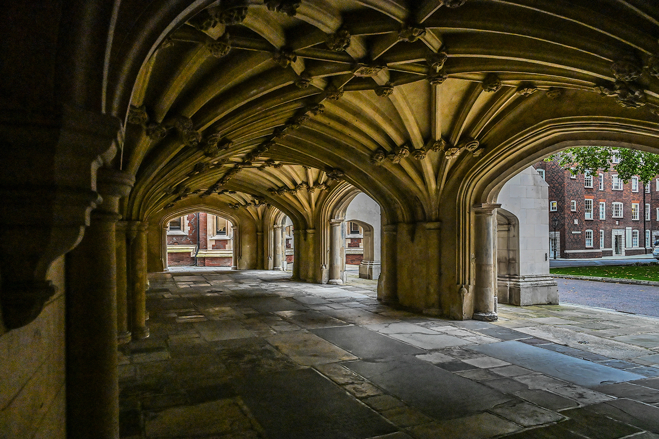 The image depicts a series of stone arches and columns forming a covered walkway, likely part of a historic or academic building. The architecture features ribbed vaults and detailed stonework, creating a sense of grandeur and antiquity. The walkway leads to an open courtyard with brick buildings and greenery visible in the background. The ground is made of large, uneven stone slabs, adding to the historic feel of the scene.