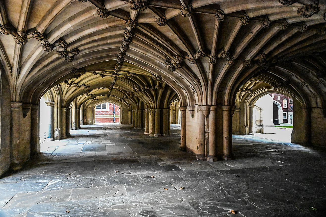 The image depicts a series of stone arches and columns forming a covered walkway or cloister. The architecture is Gothic, characterized by pointed arches and intricate stone carvings. The floor is made of large, smooth stone slabs, and the area appears to be part of a historical or academic building, possibly a university or a cathedral.