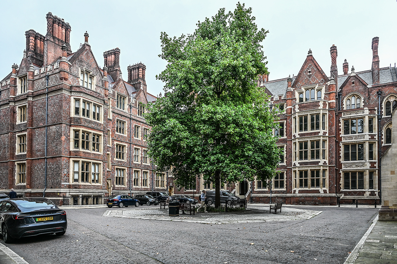 The image depicts a historic building with intricate architectural details, featuring tall, ornate windows and multiple chimneys. The building is constructed from red brick with stone accents. A large tree stands prominently in the center of the courtyard, surrounded by parked cars and a few benches. The scene is calm, with a person sitting on a bench near the tree. The overall atmosphere is serene and reminiscent of a traditional, scholarly institution.