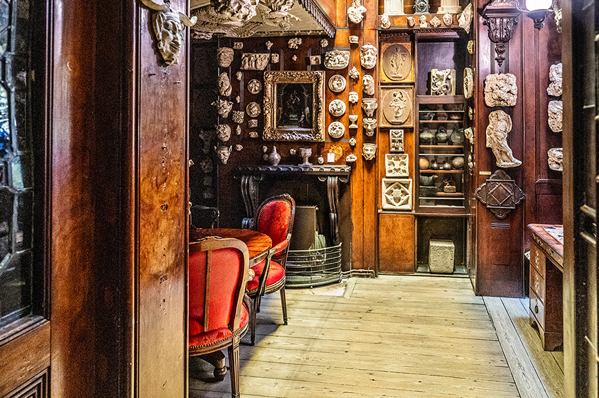 The image depicts a cozy, antique room filled with ornate wooden furniture and various decorative items. The walls are adorned with intricate carvings and framed artwork. There are two red upholstered chairs positioned near a fireplace, and the floor is made of polished wood. Shelves and cabinets display an array of decorative plates and sculptures. The overall ambiance is warm and inviting, reminiscent of a vintage interior design style.