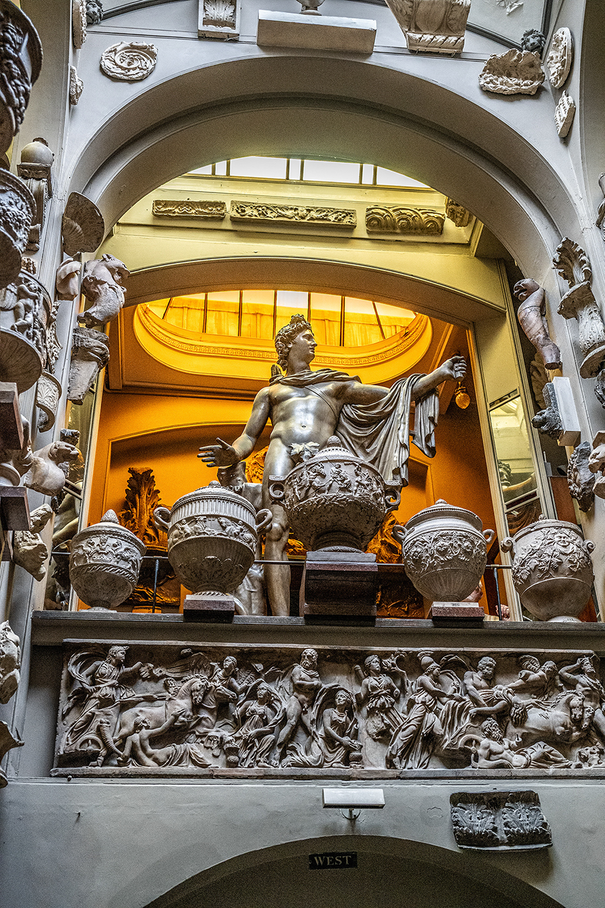 The document describes a detailed and ornate architectural scene featuring a large statue of a man holding a globe, surrounded by various decorative elements such as urns, sculptures, and intricate carvings. The setting appears to be a grand, classical interior with arches and detailed stonework.