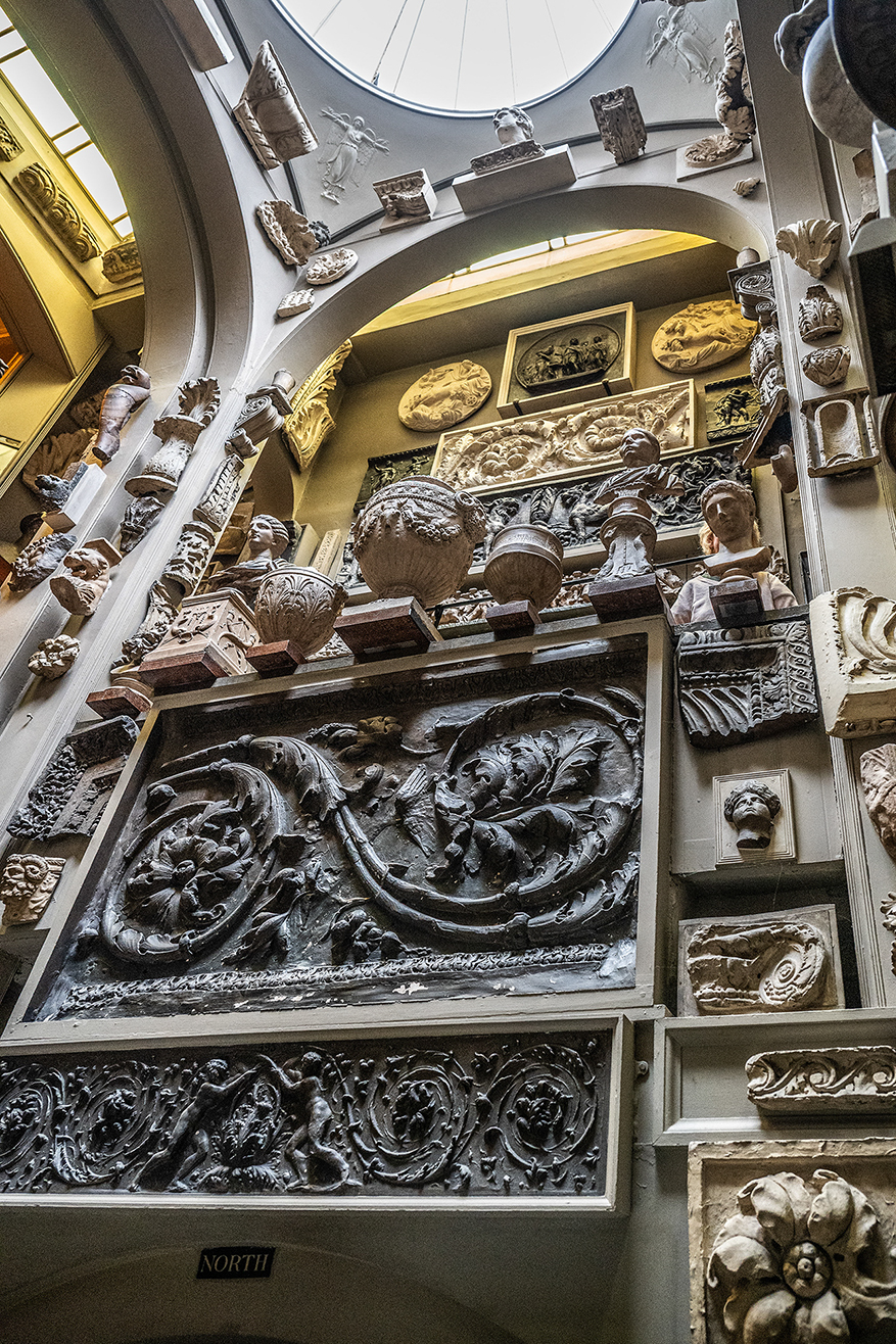 The image depicts an interior space, likely a museum or gallery, showcasing a variety of intricate and elaborate stone carvings and reliefs. The carvings include detailed depictions of figures, animals, and ornate designs. The display is organized in a structured manner, with pieces arranged on shelves and walls. The overall ambiance suggests a historical or classical theme, emphasizing the artistry and craftsmanship of stone carving.