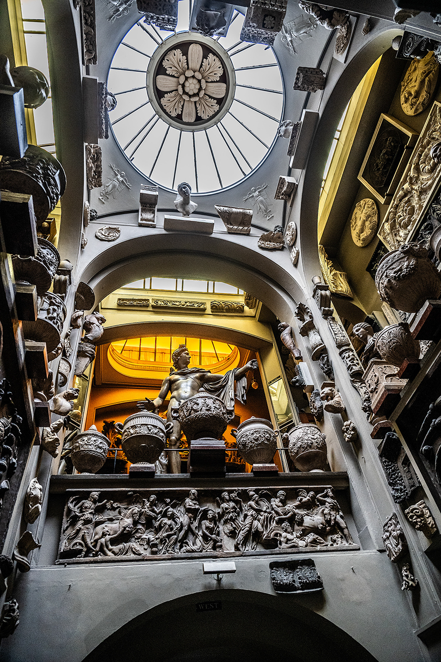 The image depicts an ornate interior space, likely a museum or gallery, featuring classical sculptures and intricate architectural details. The ceiling has a large, decorative skylight surrounded by sculpted reliefs and statues. The central statue appears to be of a classical figure, possibly a god or mythological character, standing on a pedestal adorned with elaborate carvings. The walls are lined with various sculptures and reliefs, showcasing detailed craftsmanship and artistic design.