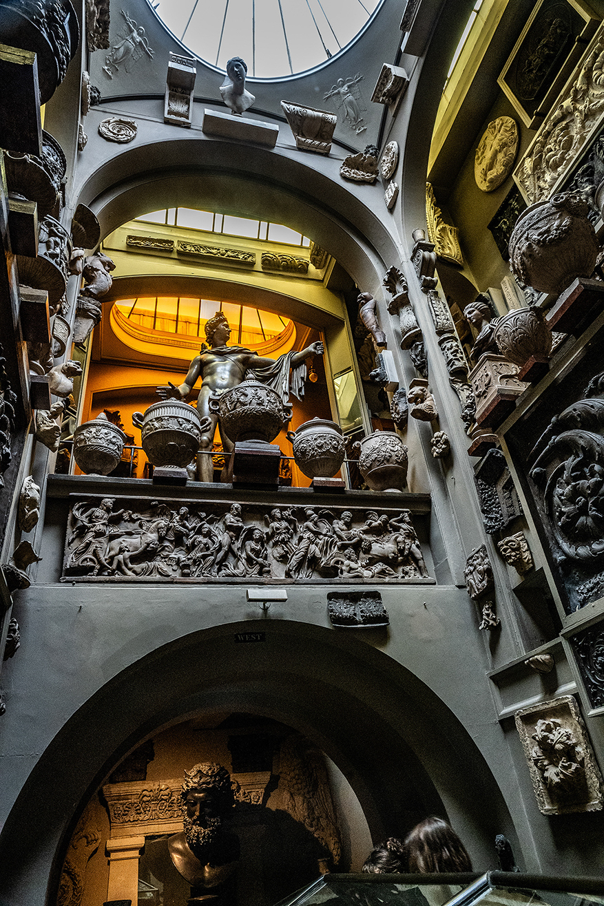 The image depicts an ornate interior space, likely a museum or gallery, featuring classical sculptures and architectural elements. The space is characterized by arched doorways, intricate carvings, and a variety of statues, including a prominent statue of a male figure holding a large urn. The ceiling is adorned with a skylight, allowing natural light to illuminate the area. The walls are decorated with detailed reliefs and various sculptural pieces, contributing to the classical and historical ambiance of the space.