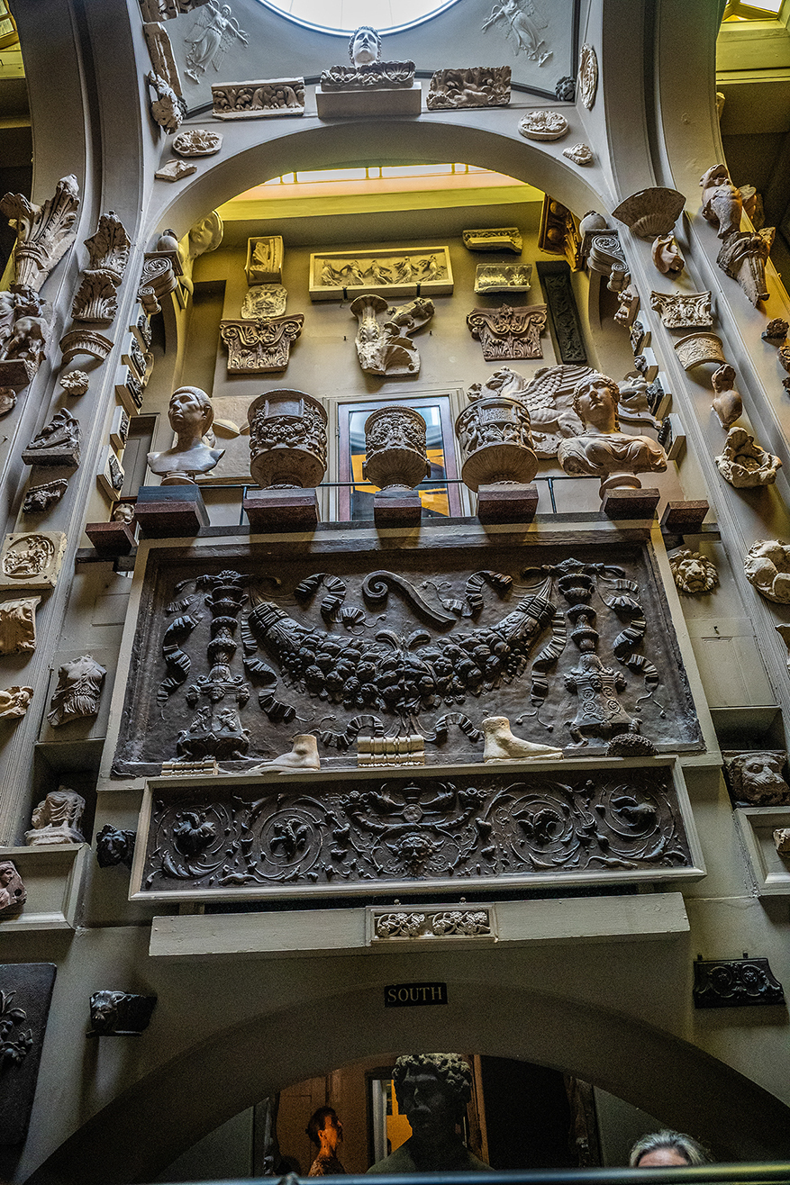 The image depicts an ornate interior space, likely a museum or gallery, showcasing a variety of classical stone carvings and sculptures. The display includes busts, decorative reliefs, and architectural elements such as columns and friezes. The central focus is a large, intricate relief panel with detailed figures and scenes, possibly from ancient mythology or history. The area is labeled 'SOUTH,' indicating a specific section or orientation within the space.