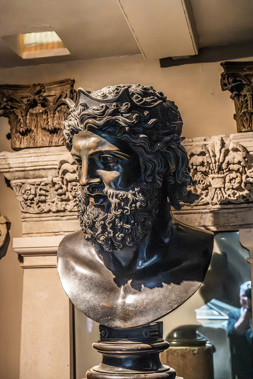 The image shows a detailed bronze bust of a bearded man with curly hair and a serious expression. The bust is mounted on a pedestal and is displayed in a museum-like setting with ornate architectural elements in the background.