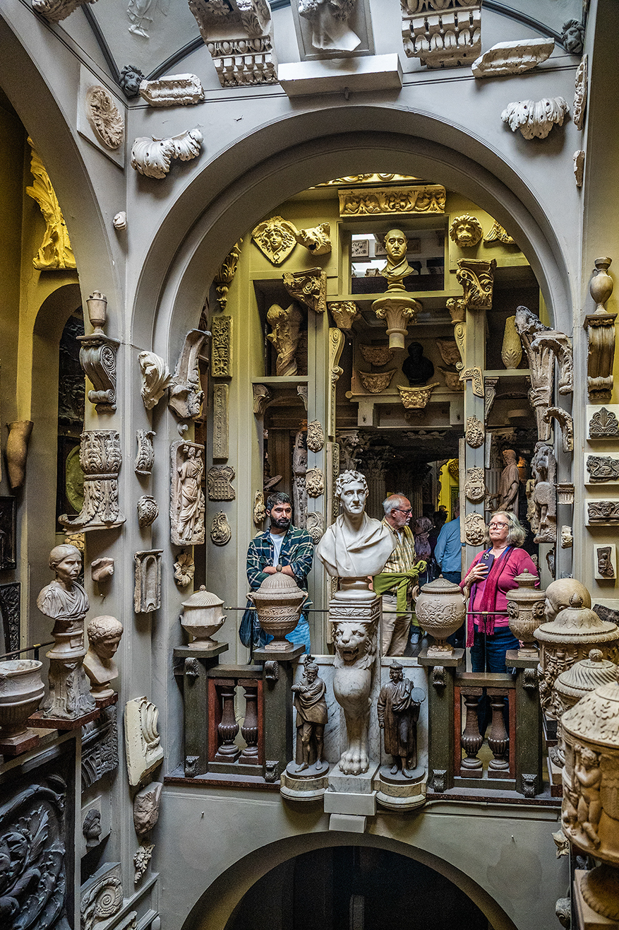 The image depicts an ornate room filled with intricate sculptures and carvings. The room features a variety of detailed reliefs and statues, including a prominent circular relief at the top center showing a dynamic scene, possibly from mythology. Below it, there are additional reliefs and sculptures, each showcasing different scenes and figures. The room is adorned with elaborate decorations, including ornate moldings and decorative elements on the walls and ceiling.