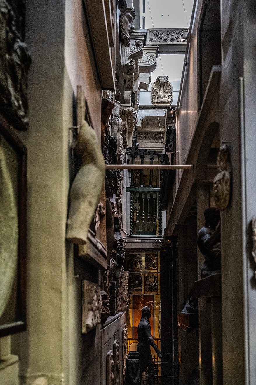 The image depicts a narrow, dimly lit alleyway with ornate architectural details and statues lining both sides. The alleyway extends into the distance, creating a sense of depth and leading to a brightly lit area at the end where a person is visible. The overall atmosphere is somewhat mysterious and intriguing.