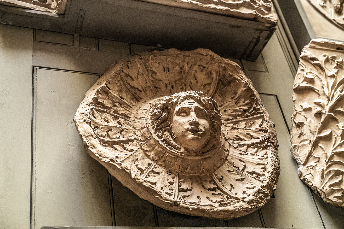 The image depicts a stone carving of a face surrounded by a sunburst pattern, likely part of an architectural element such as a keystone or decorative relief. The carving appears to be part of an older building, showcasing intricate details and weathered texture, indicative of historical or classical architecture.