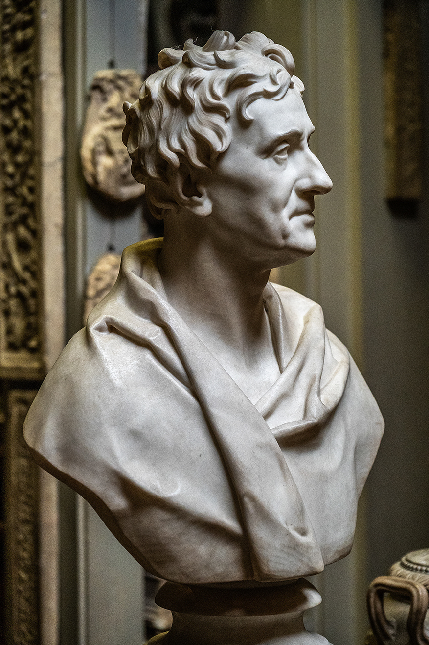 The image depicts a marble bust of a man with curly hair, wearing a draped garment. The bust is displayed in what appears to be a museum or gallery setting, with other classical art pieces visible in the background. The sculpture captures the man's likeness with detailed attention to his facial features and the texture of his hair and clothing.