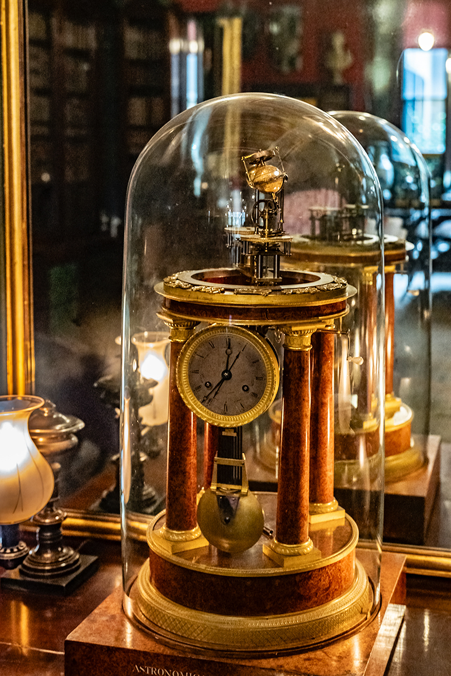 The image shows an antique clock encased in a glass dome. The clock has a wooden base with gold accents and a round face with Roman numerals. Above the clock face, there is a mechanism with a pendulum and a small golden figure that likely chimes on the hour. The clock is part of a collection, as other similar clocks are visible in the background