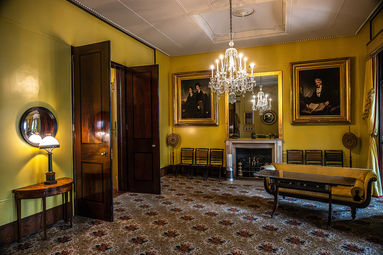 The image depicts a classic, elegantly furnished room with yellow walls, featuring a chandelier, a fireplace, and several pieces of antique furniture, including a bench and chairs. The room also has two large paintings, a round mirror, and a small table with a lamp. The overall ambiance is warm and inviting, with a touch of historical charm.