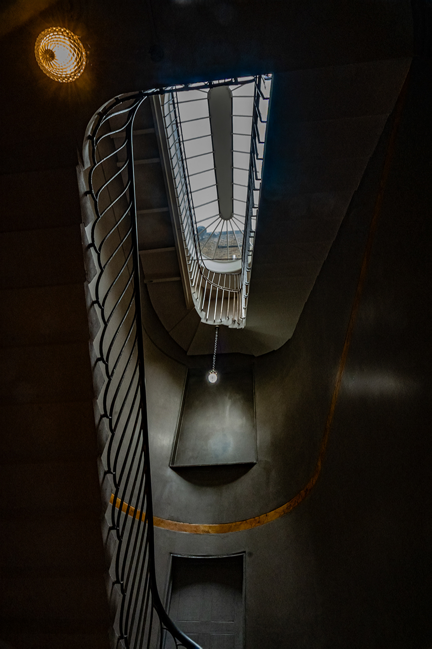 The image depicts a modern, spiral staircase with a glass railing and a light well that allows natural light to illuminate the space. The staircase is surrounded by dark walls, and there is a single light fixture hanging from the ceiling. The design is sleek and contemporary, with a focus on geometric shapes and clean lines.