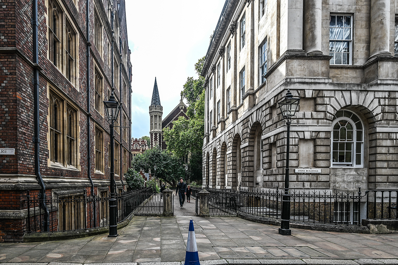 he image depicts a narrow, paved pathway flanked by historic buildings on both sides. The architecture features a mix of brick and stone, with large windows and decorative elements. Traditional street lamps line the pathway, and a few people are walking in the distance. The pathway leads to a building with a prominent spire in the background. The overall atmosphere is serene and reminiscent of a university or institutional setting.