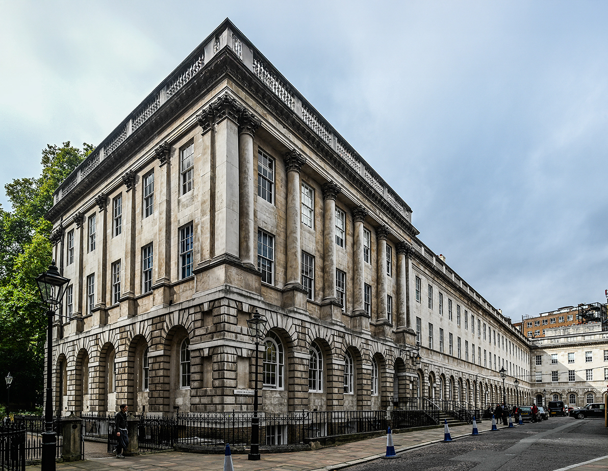 The image depicts a grand, historic building with classical architectural elements such as columns and arched windows. The building is part of a row of similar structures, likely in a city setting. The facade is made of stone, and the overall style suggests it could be a significant public or institutional building, possibly in a European city.