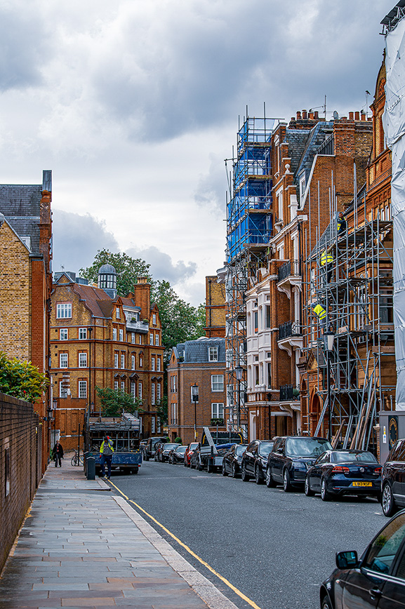 The image depicts a street in a residential area with brick buildings, some of which are under construction or renovation as evidenced by the scaffolding. Cars are parked along the street, and a few people are visible walking or riding bicycles. The sky is overcast, suggesting it might rain soon.