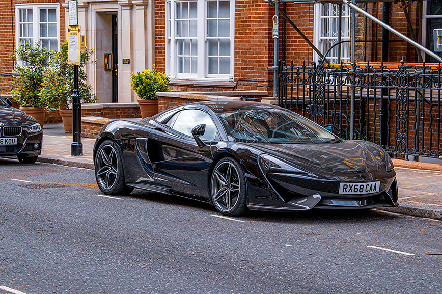 The image shows a sleek, black sports car parked on a street in front of a brick building with large windows and a black wrought iron fence. The car has a low profile, aerodynamic design, and distinctive features such as a large air intake on the side and a prominent front splitter. The license plate reads 'RX68 CAA'. There is another car parked behind the sports car, and the street is lined with potted plants and a lamppost.