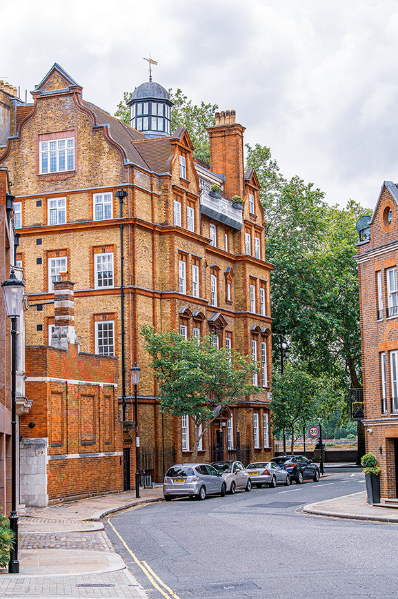 The image depicts a street scene with a prominent, multi-story brick building featuring classic architectural details such as arched windows, decorative cornices, and a dome-like structure on the roof. The building has a distinct, historical appearance with a mix of red and brown bricks. Several cars are parked along the street in front of the building, and the street itself is lined with trees and traditional street lamps. The overall setting suggests a quiet, possibly residential or upscale urban area.