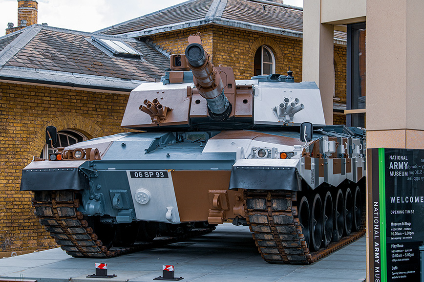 The image shows a military tank displayed outside the National Army Museum. The tank has the identification number '06 SP 93' and features camouflage paint. The museum's sign is visible, providing information about opening times and facilities. The setting appears to be an urban area with brick buildings in the background.