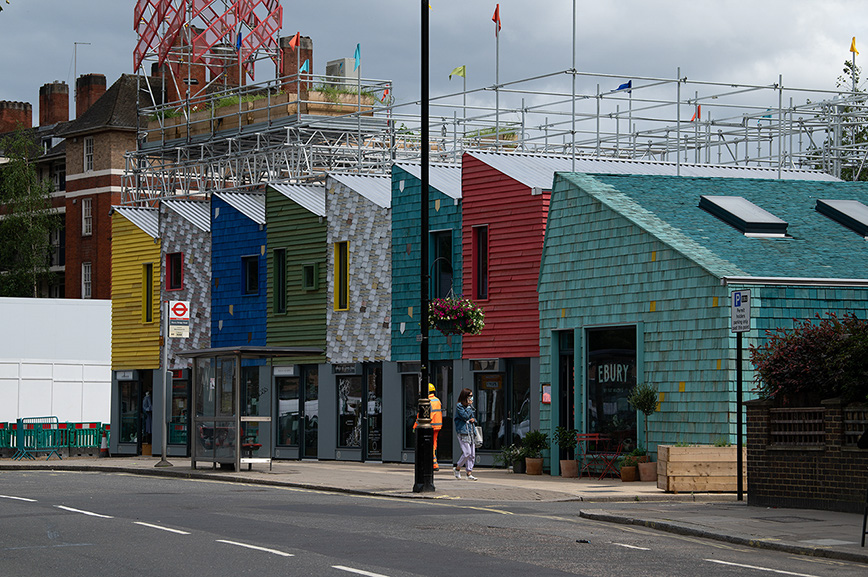 The image depicts a row of colorful buildings with various architectural styles and facades, including a yellow, blue, green, red, and teal structure. The buildings are part of a commercial area with shops and businesses. There are construction scaffolding and structures visible on top of the buildings, indicating ongoing construction or renovation work. The street in front of the buildings is relatively quiet, with a few pedestrians and a traffic sign visible. The overall scene suggests a vibrant and developing commercial district.