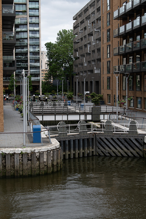 The image depicts a modern urban waterfront scene with high-rise residential buildings, a pedestrian walkway, and a dock area adjacent to a body of water. The buildings feature balconies and a mix of brick and glass exteriors. The dock area includes railings, lighting fixtures, and some planters with greenery.