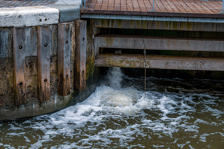 The image shows a section of a waterway with a small waterfall or spillway. Water is flowing over a wooden structure into a lower body of water. The structure appears to be part of a lock or dam, with visible bolts and a chain hanging down. The surroundings include a concrete or stone wall and a wooden deck or walkway above.