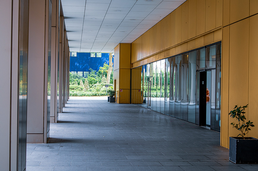 The image depicts a modern, open corridor with a tiled floor and large glass windows on one side. The corridor is lined with wooden panels on the ceiling and part of the walls. There are a few doors along the corridor, and a potted plant is visible on the right side near the end of the corridor. The area outside the windows appears to be a landscaped garden with greenery and a blue structure in the background.