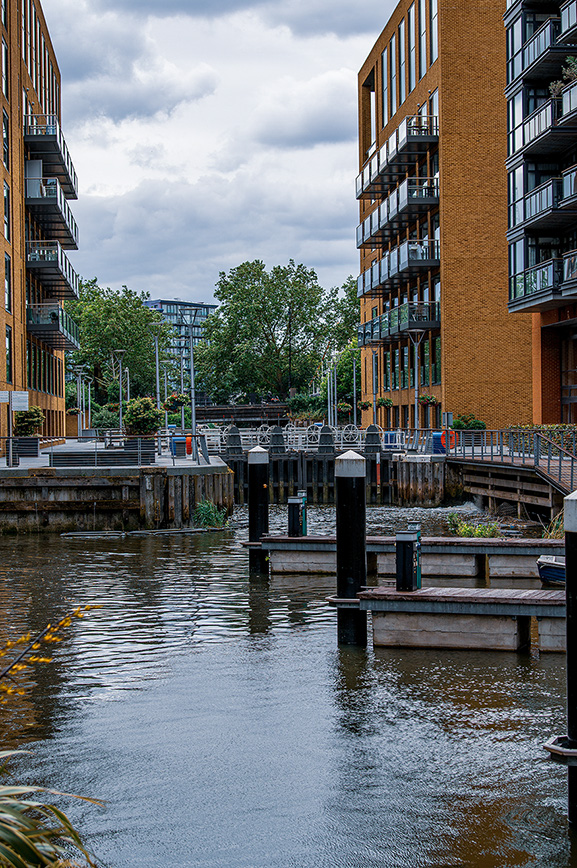 The image depicts a serene canal flanked by modern residential buildings with balconies. The waterway is bordered by walkways and greenery, with a bridge in the background connecting the two sides. The overall atmosphere is calm and urban, suggesting a peaceful residential area.