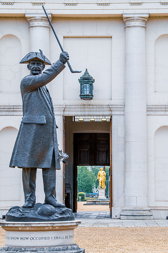 The image shows a statue of a man in historical attire, standing in front of a building with columns. The statue holds a pole in one hand and has a tricorn hat. The base of the statue has an inscription. Through the open doors of the building, another statue can be seen in the background.
